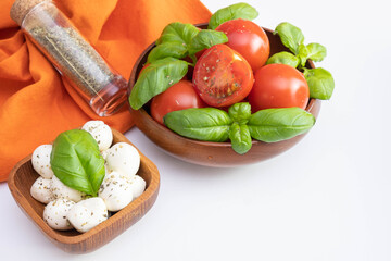 fresh tomatoes and green basil in a wooden bowl, next to a small mozzarella in a wooden bowl and spices on a white table