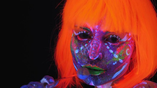 Portrait of a beautiful girl with fluorescent makeup and a bright orange wig in the Studio with a Lollipop on a black background.