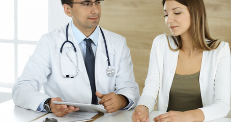 Obraz premium Doctor and patient discussing medical exam results while sitting at the desk in clinic. Male physician using tablet computer for filling up medication history record of young woman. Data in medicine