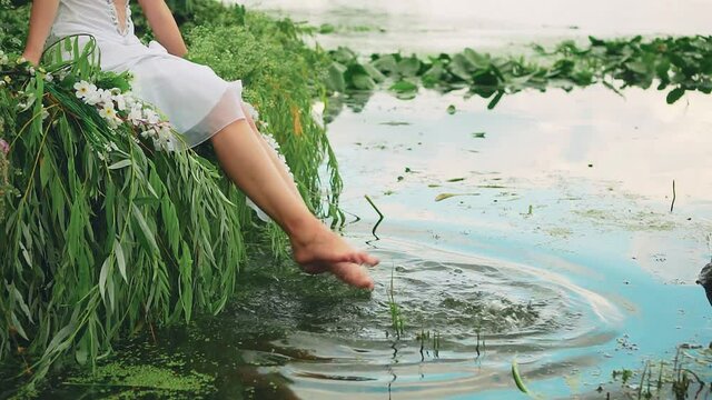 Women's feet splash in the river water. Happy girl in white dress sits on boat, beautiful long naked legs. Summer green nature background, water, lake, trees, water lily flowers. Fantasy woman nymph