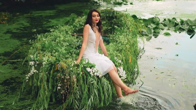 Cheerful woman in a white vintage outfit sits on a boat and splashes her bare feet in the water. wild background river with water lilies, summer green trees. Happy girl Princess bide in wedding dress.