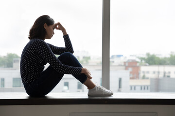 Depressed millennial korean mixed race woman sitting on windowsill crying regretting relations...