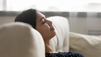 Close up head shot happy serene millennial asian mixed race woman leaning on comfortable sofa, daydreaming sleeping napping restoring energy, enjoying tranquil peaceful weekend time at home.