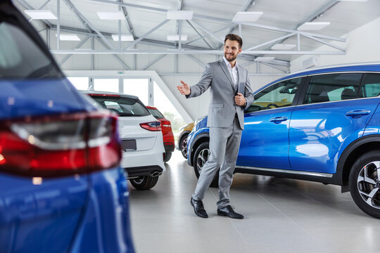 Smiling Friendly Car Seller In Suit Walking Around Car Salon And Showing Around Cars.