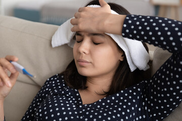 Unhealthy young korean asian woman measuring body temperature with electronic thermometer, applying wet towel compress to forehead relieving strong headache, seasonal contagious grippe illness concept