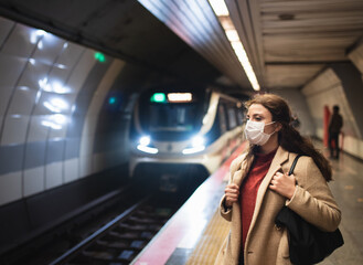 Beautiful girl wearing protective medical mask and fashionable clothes waits for train on subway platform