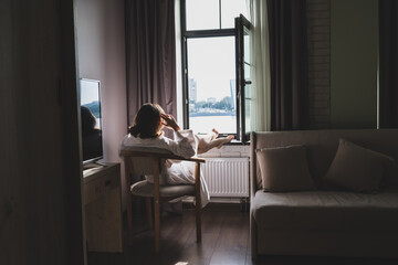 Back view of beautiful young woman in bathrobe sitting on armchair and enjoying near open window at sunny morning.