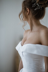 A bride in a beautiful white dress stands with her back at the light window.