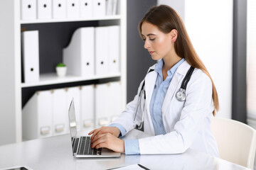 Doctor woman typing on laptop computer while sitting at the desk in hospital office. Physician at work. Medicine and health care concept