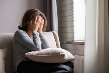 Sad scared woman covering her face with hands and crying. Lonely girl sitting near window with pillow on a sofa of apartments.