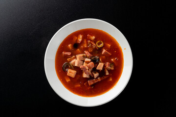 Hot gourmet meat soup solyanka with black olives and tomato broth, bright red on black
