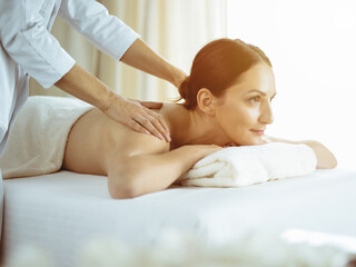 Obraz na płótnie Canvas Pretty brunette woman enjoying procedure of back massage in sunny spa salon. Beauty concept. Cold toned picture