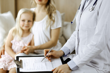 Doctor and patient. Pediatrician using clipboard while examining little girl with her mother at home. Happy cute caucasian child at medical exam. Medicine concept