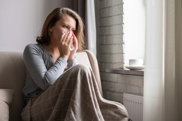 Sick brunette woman sneezing in a tissue sitting on the couch in the living room near window