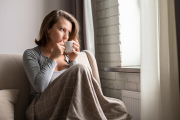 Portrait of beautiful сaucasian young woman sitting on sofa covered blanket, drinking hot coffee from cup and looking at window.
