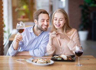 Affectionate millennial couple taking photo of yummy food and wine on smartphone at restaurant