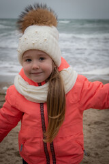 a little girl in a winter suit plays on the seashore, a girl poses on the seashore, a little girl in a hat with a bumbon plays near the sea