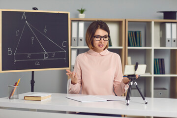 Female math and geometry teacher explains new material to students through a video call using a phone on a tripod. Woman sits in a classroom with a blackboard next to her and conducts an online lesson