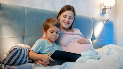 Family lying in bed and watching cartoons on tablet computer at night. Little boy and pregnant mother using digital tablet in bed at night