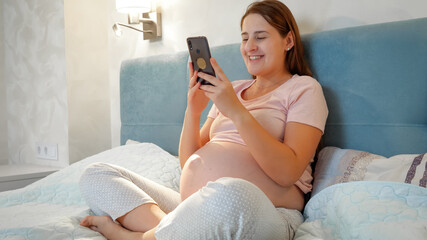 Young pregnant woman talking on video call while sitting on bed at night. Pregnant girl talking to...