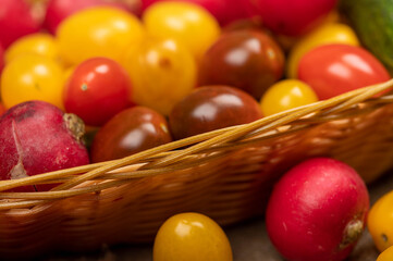 Green cucumbers, ripe radishes and colorful tomatoes in a wicker basket and scattered on the table. Close-up, selective focus.
