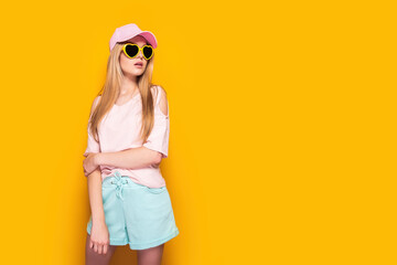 Elegant blonde woman wearing fashion cap, sunglasses enjoying sun on yellow summer background.