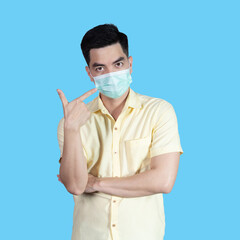 COVID-19 Coronavirus portrait handsome young asian man wearing yellow shirt and mask protection from covid 19 and thumbs up sign isolated on blue background in studio. Asian people. COVID-19 concept.
