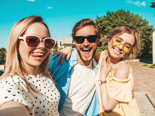 Group of young three stylish friends in the street.Man and two cute female dressed in casual summer clothes.Smiling models having fun in sunglasses.Women and guy making photo selfie on smartphone