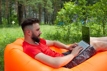 Obraz premium Bearded man is working and smiling with laptop at the park under the tree. Happy freelancer is sitting and using app or website on the grass. Work remotely in self-isolation mode