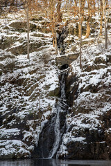 Small waterfall with a path and stone bridge on a hillside. A layer of snow frames the scene