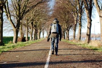 A man walking on a road with a backpack with a row of trees and green meadow. Outdoor hiking activities for healthy living concept.
