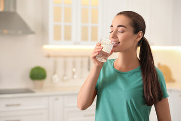 Young woman drinking pure water from glass in kitchen. Space for text