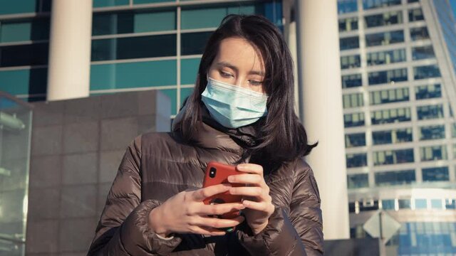 Young woman in protective mask with phone, outside on background of business buildings. Girl in medical protective mask in the city center outdoors, sunny autumn weather
