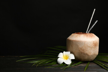 Obraz na płótnie Canvas Fresh coconut with drinking straws and flower on wooden table against black background. Space for text