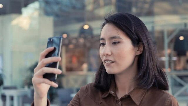 Young woman with phone making selfie on background of glass windows in city mall. Asian girl filming herself standing in business city mall