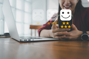 Customer service evaluation concept. smiling Asian female Is using a smartphone And she is pressing face emoticon smiling in satisfaction on virtual touch screen. - obrazy, fototapety, plakaty