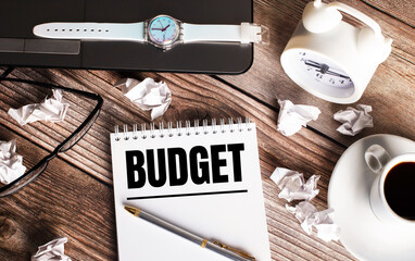 There is a cup of coffee on a wooden table, a clock, glasses and a notebook with the word BUDGET. Business concept