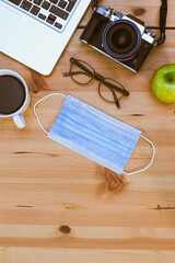 Camera, a laptop computer, an apple, a facial medical mask and a coffee cup filled with coffee on a wooden background.