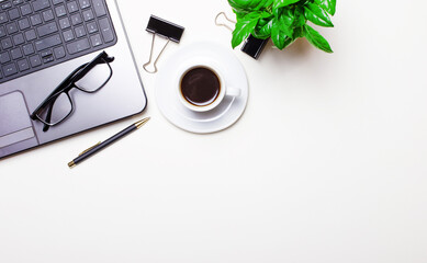 On a white desktop, a laptop, glasses, a pen, coffee and a plant in a pot. Top view with copy space