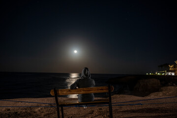 Plakat woman sits on a bench and looks at the rising moon over the sea at night