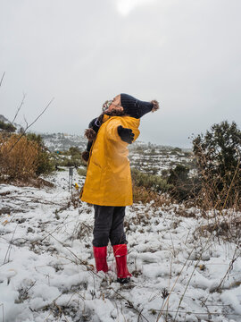 A girl in a yellow jacket and red boots playing happily in the snow with her arms raised