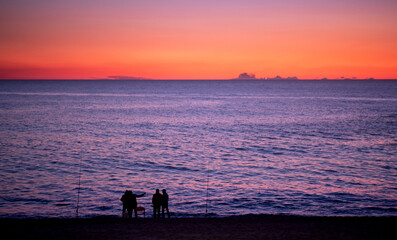 Fishermen in silhouette on beach at dusk with vivid red sky, fishing in the ocean.