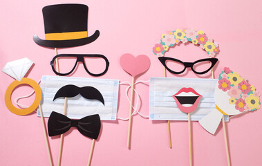 Fototapeta premium Paper groom and bride consisting of sunglasses, hat, mustache, lips with a medical mask, bridal bouquet, wedding ring on the pink background. Quarantine wedding party. Funny fashion.