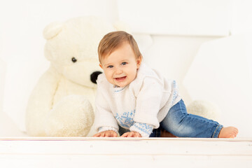 a happy little child six months old in a knitted warm jacket and blue jeans crawls at home near a large Teddy bear