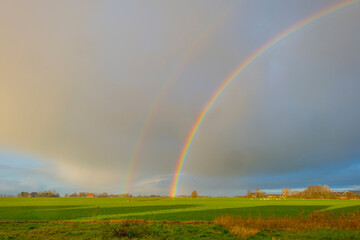 Double rainbow and yellow grey rain clouds over a windy rainy green meadow in bright sunlight in winter, Leeuwarden, Friesland, The Netherlands, January 9, 2021