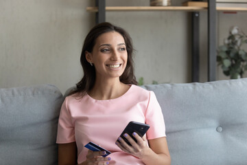 Smiling millennial indian Arabic woman sit on sofa at home shopping online on cellphone with credit card. Happy young ethnic female client buyer pay on internet use secure bank system on smartphone.