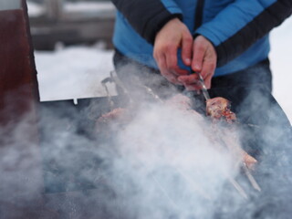Hands of a man fry on the street in winter, pieces of bird on skewers on coals and an open fire.Blurred image from steam and smoke.