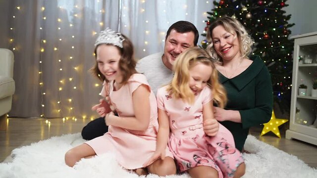 A cheerful happy family sits on the floor of the house before the Christmas tree fools around and laughs. Dad, mom and two daughters at a New Year's Eve party at home.