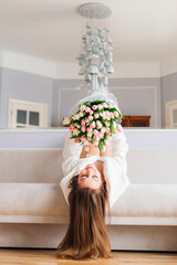 Happy young woman lying on sofa and holding a large bouquet of tulips over her at home. The bride is lying on a white bed and hold large flowers in the hotel.
