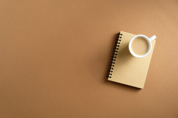 Cup of coffee and paper notepad on brown background top view. Cozy home office desk, elegant feminine workspace.